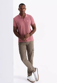 Camisa polo rosa de manga corta con un pequeño emblema, combinada con pantalones cargo beiges que tienen bolsillos laterales y zapatillas blancas.