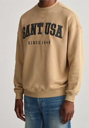 Beige sweatshirt med teksten "GANT USA SINCE 1949" båret med blå jeans, vist fra skuldre til hofter, neutral baggrund.