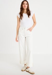 Mujer joven con cabello largo y castaño, vistiendo una blusa blanca ajustada sin mangas, pantalones blancos de pierna ancha y zapatillas blancas, de pie con una mano en el bolsillo.