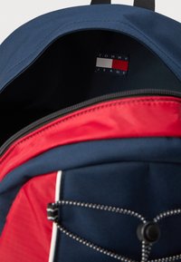 Námornícky modrý a červený látkový batoh s horným zipsom, ktorý má vo vnútri štítok Tommy Jeans a dekoratívny detail so stuhou na vonkajšej strane.