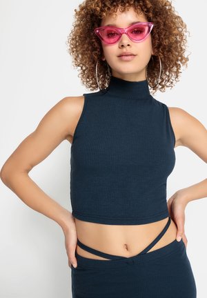Navyblaue gerippte Cropped-Top mit hohem Kragen, ärmellosem Design und verstellbarem Trägerdetail in der Taille. Getragen mit pinken Katzenaugen-Sonnenbrillen.