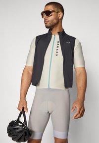 Vêtements de cyclisme avec un gilet sans manches noir sur un maillot à manches courtes beige clair, un short rembourré gris et un casque noir.