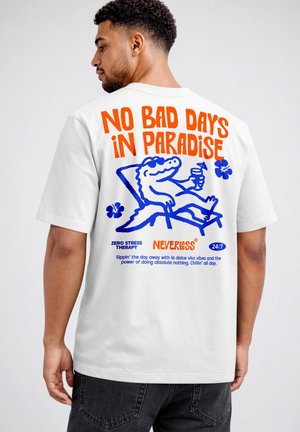 Man wearing white T-shirt with colorful text "No bad days in paradise" and cartoon alligator lounging with a drink on the back.