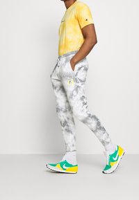 Man som bär en gul batikskjorta, grå och vit batik-joggingbyxor samt gröna, gula och vita sneakers, går mot en vit bakgrund.