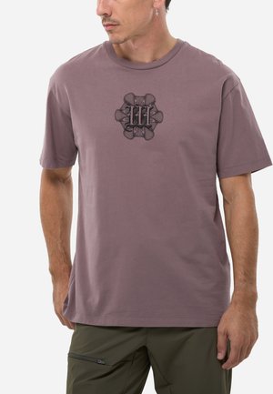 Homme portant un t-shirt manches courtes mauve avec un motif orné en forme de lettre "M" noire, un pantalon vert olive avec une poche zippée, et une boucle d'oreille en forme de croix.