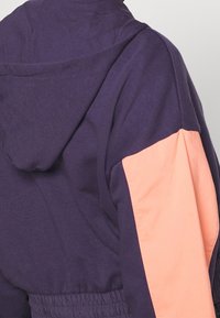 Fialová mikina s kapucí se korálovým panelem na levém rukávu. Měkká látka s elastickým pasem, volný střih a žádné viditelné kovové prvky.