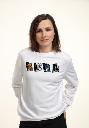 Kobieta o brązowych, sięgających ramion włosach, ubrana w białą bluzę z stylizowanymi hełmami oznaczonymi jako Armorer, Mando, Paz Vizsla i Bo-Katan.