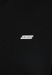 Czarna koszulka z prążkowanej tkaniny z małą białą metką logo "SMILODOX" z przodu. Prosty design, dopasowany krój.