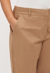 Tan bukser med en glat, tekstureret stof. Har flad front med sidelommer og en slim fit. Rene linjer uden synlig hardware.