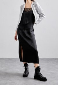 Femme portant une robe de nuit en satin noir avec des dentelles, une veste en tricot gris et des bottes de combat noires à lacets sur un sol gris.