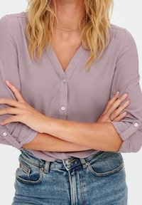 Blusa di lavanda chiara con scollo a V, chiusura con bottoni e maniche arrotolate. Abbinata a jeans in denim a vita alta. Dettaglio del tessuto testurizzato visibile.