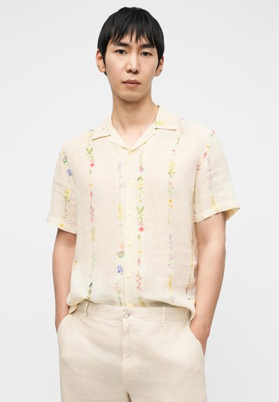 Κοντομάνικο πουκάμισο από κρεμ λινό με πολύχρωμη κεντητή λουλουδένια διακόσμηση, διαθέτει κουμπιά στο μπροστινό μέρος και φαρδιά εφαρμογή. Συνδυάζεται με ανοιχτόχρωμο παντελόνι.