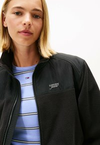 Giacca in pile nera con zip frontale, caratterizzata da un logo sul petto e abbinata a una maglietta a righe blu chiaro sotto.