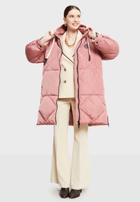 Chaqueta rosa acolchada con capucha y ribete negro, con parche de logo y bolsillos laterales. Llevada sobre un traje de color claro.