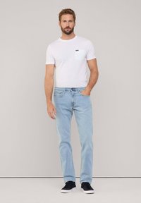 Camiseta blanca de manga corta con bolsillo, combinada con jeans de corte claro. El modelo lleva zapatillas negras. Diseño sencillo, corte informal.