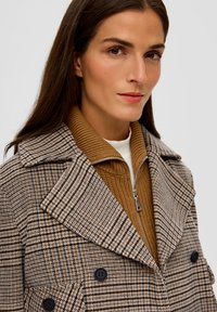 Płaszcz w kratę w odcieniach beżu i brązu z wełny, z szerokimi klapami, podwójnym zapięciem i czarnymi guzikami, noszony na beżowym swetrze robionym na drutach.