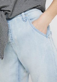 Světle modré džíny s uvolněným střihem, přední kapsou a jemnou texturou. Spárovány s topem v černo-bílém kostkovaném vzoru.