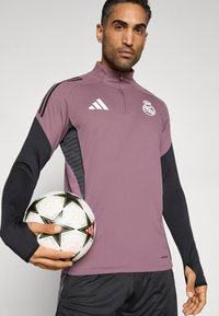 Αθλητικό μακρυμάνικο μπλουζάκι επόμενης γενιάς σε μωβ απόχρωση με μαύρες λεπτομέρειες, διαθέτει όρθιο γιακά, φερμουάρ και λογότυπο συλλόγου· συνδυασμένο με ποδοσφαιρικό μπάλα.