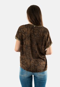 Camisa marrón de manga corta con un patrón de manchas y ribete verde, que presenta un dobladillo ligeramente redondeado y un pequeño logo en la parte posterior.