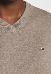 V-Ausschnitt-Pullover aus hellbraunem Strickmaterial, mit geripptem Ausschnitt und einem kleinen Logo-Akzent in Rot, Weiß und Blau.