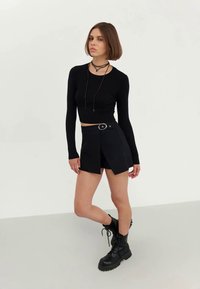 Haut noir à manches longues à côtes associé à une jupe portefeuille noire avec une grande boucle circulaire, complété par des bottines militaire noires à lacets.