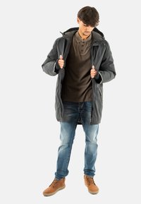 Chaqueta acolchada gris oscuro con capucha, cierre frontal con cremallera y puños ajustables, llevada sobre una camiseta polo marrón y pantalones vaqueros azules. Zapatos marrones.