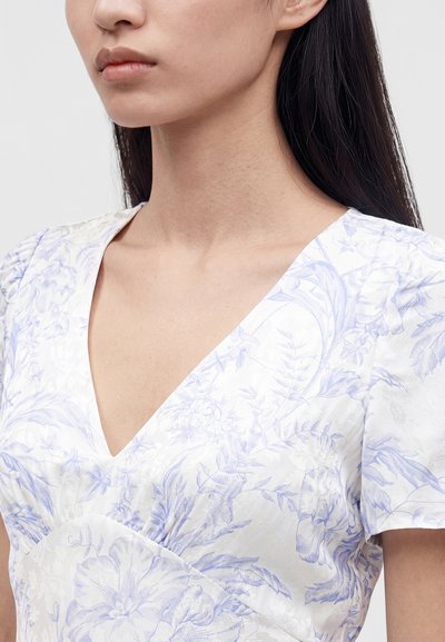 Femme portant une robe blanche avec des motifs floraux bleu clair et un profond décolleté en V, montrant son cou et une partie de son visage et de son épaule.