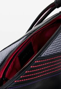 Bolso de cuero negro con cierre de cremallera; el interior presenta un forro rojo, un bolsillo texturizado gris y costuras vibrantes en rosa y púrpura.