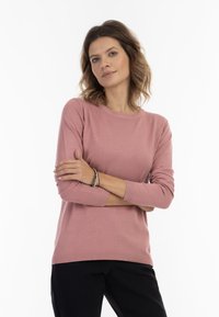Lichtroze gebreide trui met een ronde halslijn en driekwart mouwen, met een geribde zoom. In combinatie met zwarte broek.