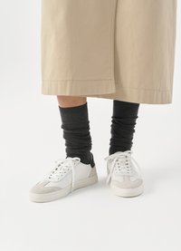 Zapatillas blancas y grises con un acento negro en el talón, con una parte superior de cuero suave y una punta de ante texturizado. Llevadas con calcetas oscuras y holgadas.
