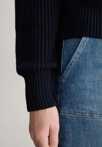 Granatowy sweter z prążkowanej dzianiny o teksturowanym wzorze, z grubym mankietem. Połączony z jasnoniebieskimi dżinsami, ukazującymi detal kieszeni.