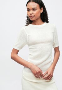 Γυναίκα με σγουρά μαλλιά, φορώντας ένα λευκό πλεκτό τοπ κοντό μανίκι και μια ριμπ λευκή φούστα, που στέκεται μπροστά σε μια απλή ανοιχτόχρωμη φόντο.