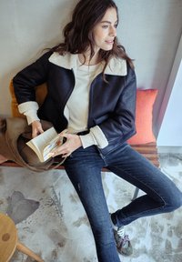 Mustat tekonahkainen takki, jossa on kermaisen vaaleat shearling-kaulus, yhdistetty valkoiseen yläosaan ja tummiin skinny-farkkuihin. Pitelee kirjaa ja istuu puupenkillä.