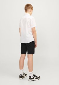 Witte korte mouwen shirt met opgerolde manchetten, gedragen met zwarte shorts. De outfit wordt aangevuld met wit en zwarte sneakers en witte sokken.