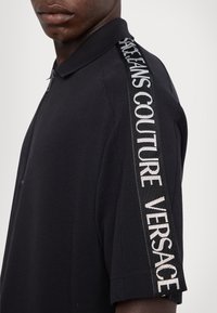 Čierna polokošeľa s textúrovanou tkaninou, krátkymi rukávmi a výraznou vertikálnou značkovou páskou s nápismi "VERSACE" a "JEANS COUTURE" v bielej farbe.