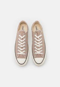 Sneakers in tela di colore marrone chiaro con lacci bianchi, punta in gomma rinforzata e linea nera lungo la suola. Design piatto, stile casual.