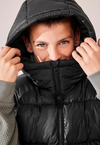 Chaleco puffer negro con un cuello alto y cierre de cremallera. El interior presenta una tela texturada gris, y el material exterior tiene un acabado brillante.