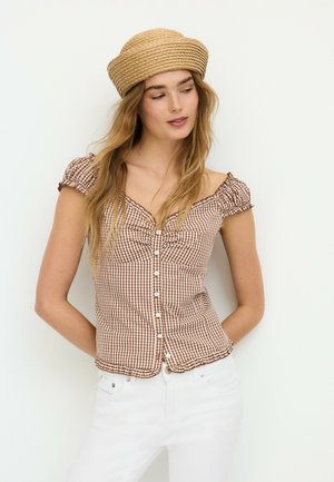 Femme portant un chemisier marron vichy à épaules dénudées, pantalon blanc et un chapeau tissé beige, regardant sur le côté devant un fond blanc uni.
