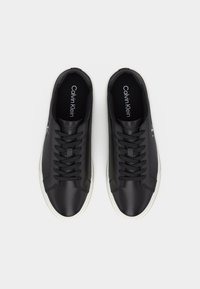 Ζευγάρι μαύρα αθλητικά Calvin Klein χαμηλού τύπου με λευκές σόλες, μαύρα κορδόνια και ορατό λογότυπο στο εσωτερικό και στο πλάι των παπουτσιών, θέα από πάνω.