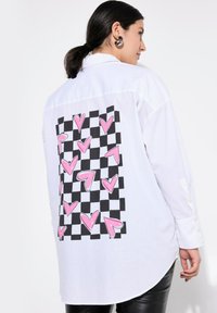 Maglietta bianca a maniche lunghe con una grafica a scacchi bianca e nera e cuori rosa sul retro. Dettagli con bottoni sulle maniche.