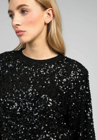 Pull en sequins noirs avec un décolleté rond, doté d'une surface texturée composée de sequins noirs et argentés brillants.