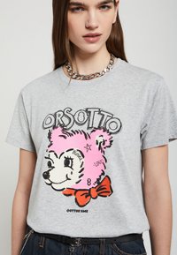 Camiseta de algodón gris con un oso de dibujos animados con pelaje rosa, contornos negros y una pajarita roja. El texto dice "ORSOTTO" y se incluyen detalles como estrellas y lágrimas.