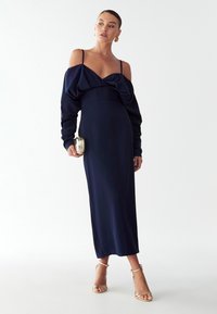 Námořnická modrá šaty bez ramínek s volánkovým korzetem, dlouhými rukávy a těsným střihem. Slučují se s páskovými sandály a malou peněženkou.