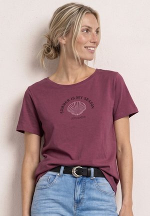 Kvinde med blondt hår sat i knold, iført en vinrød t-shirt med teksten "SUMMER IS MY SEASON CÔTE D'AZUR" og en muslingegrafik, kombineret med lyseblå jeans.