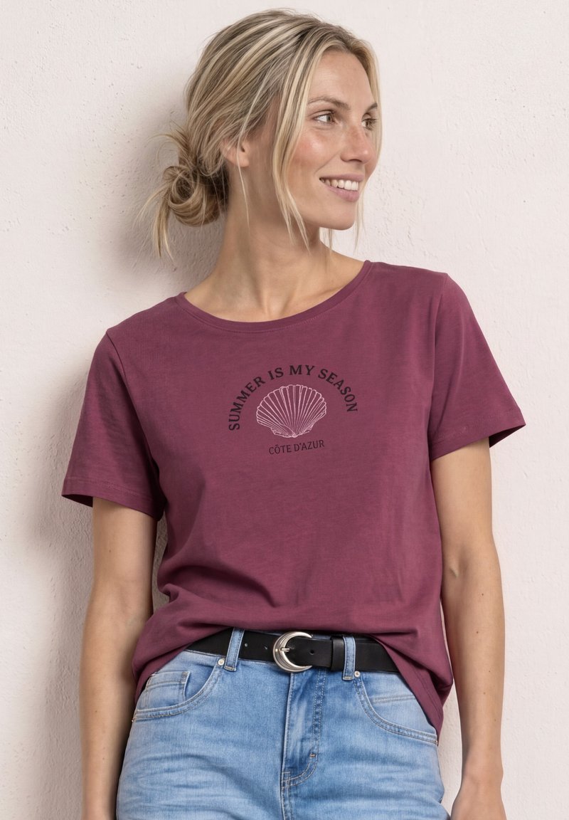 Donna con capelli biondi raccolti in uno chignon che indossa una t-shirt color bordeaux con la scritta "SUMMER IS MY SEASON CÔTE D'AZUR" e una grafica di una conchiglia, abbinata a jeans azzurri chiari.