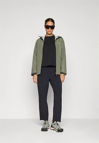 Lehká olivově zelená bunda se šedou podšívkou, doplněná černým topem a tmavými kalhotami s kapsami na zip. Šedé a černé tenisky.