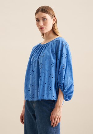 Blusa di cotone blu con maniche a palloncino, caratterizzata da motivi floreali intagliati, scollatura tonda e vestibilità rilassata. Abbinata a jeans in denim.