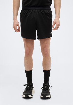 Mies, jolla on päällään mustat urheilushortsit, mustat keskipitkät sukat ja mustat juoksukengät, seisoo yksinkertaisen vaalean taustan edessä.