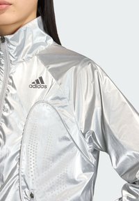 Srebrna sportowa kurtka z wysokim kołnierzem, wyposażona w odblaskowy materiał, akcenty w postaci wzoru z kropek oraz logo Adidas na lewej górnej części.