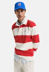 Rood-witte gestreepte lange mouw polo met kraag, over een lichtblauwe gekraagde shirt, gecombineerd met beige broek en een blauwe pet.